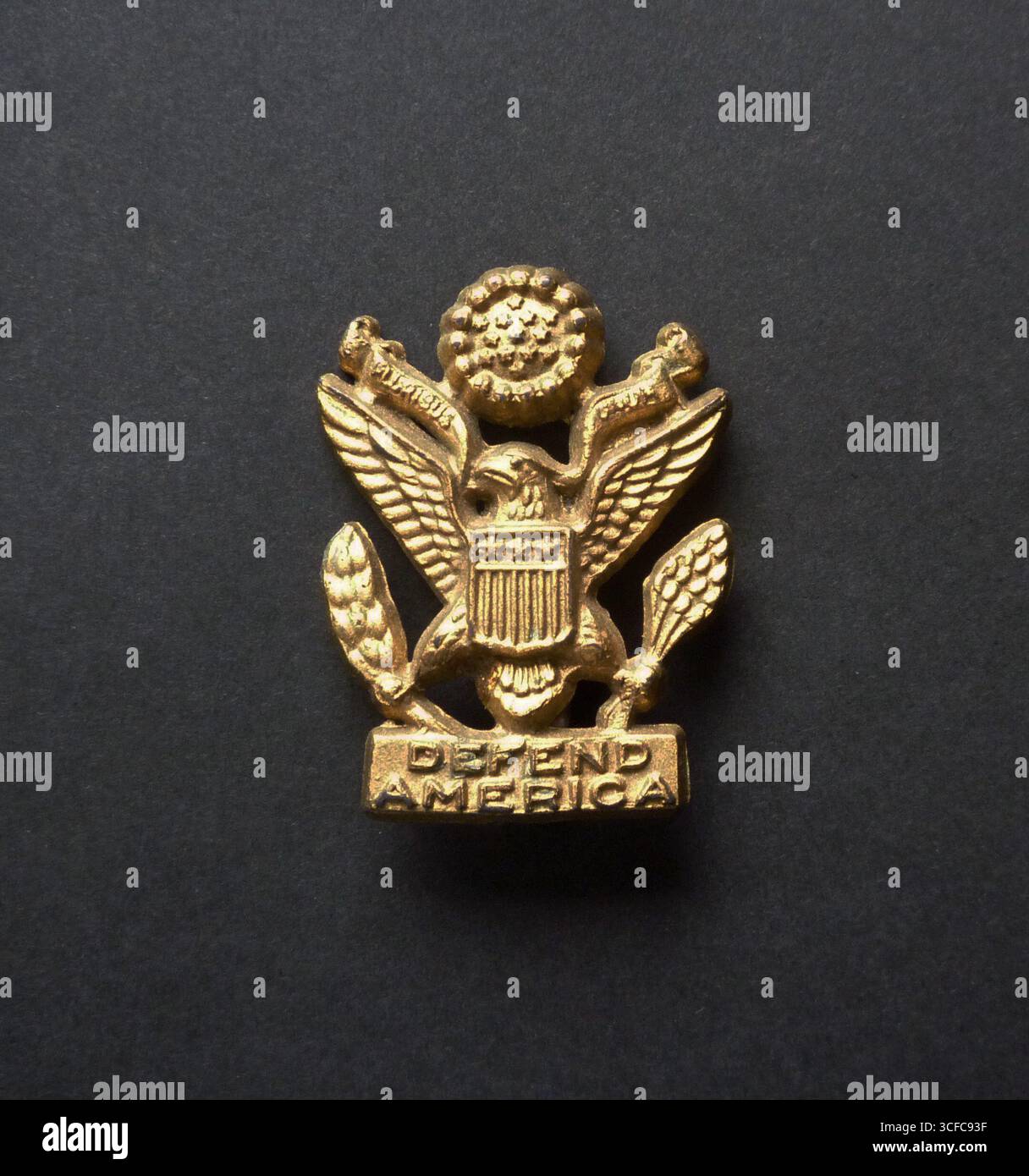Badge patriotique de la seconde Guerre mondiale en plastique peint or sous la forme du Grand Sceau des États-Unis. Ci-dessous se trouve la légende, « défendre l’Amérique ». Banque D'Images