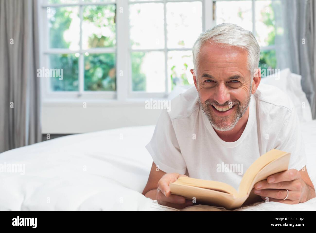 Homme senior portant T-shirt blanc couché sur le lit et lisant livre de poche dans la chambre à coucher par la fenêtre Banque D'Images