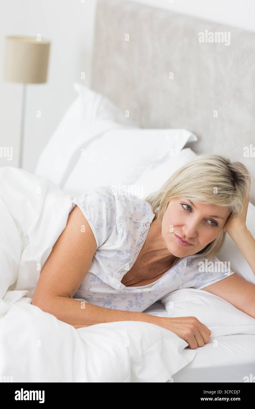 Femme mature couchée sur le lit sous la tête de lit rembourrée grise dans la chambre portant un haut de pyjama floral Banque D'Images