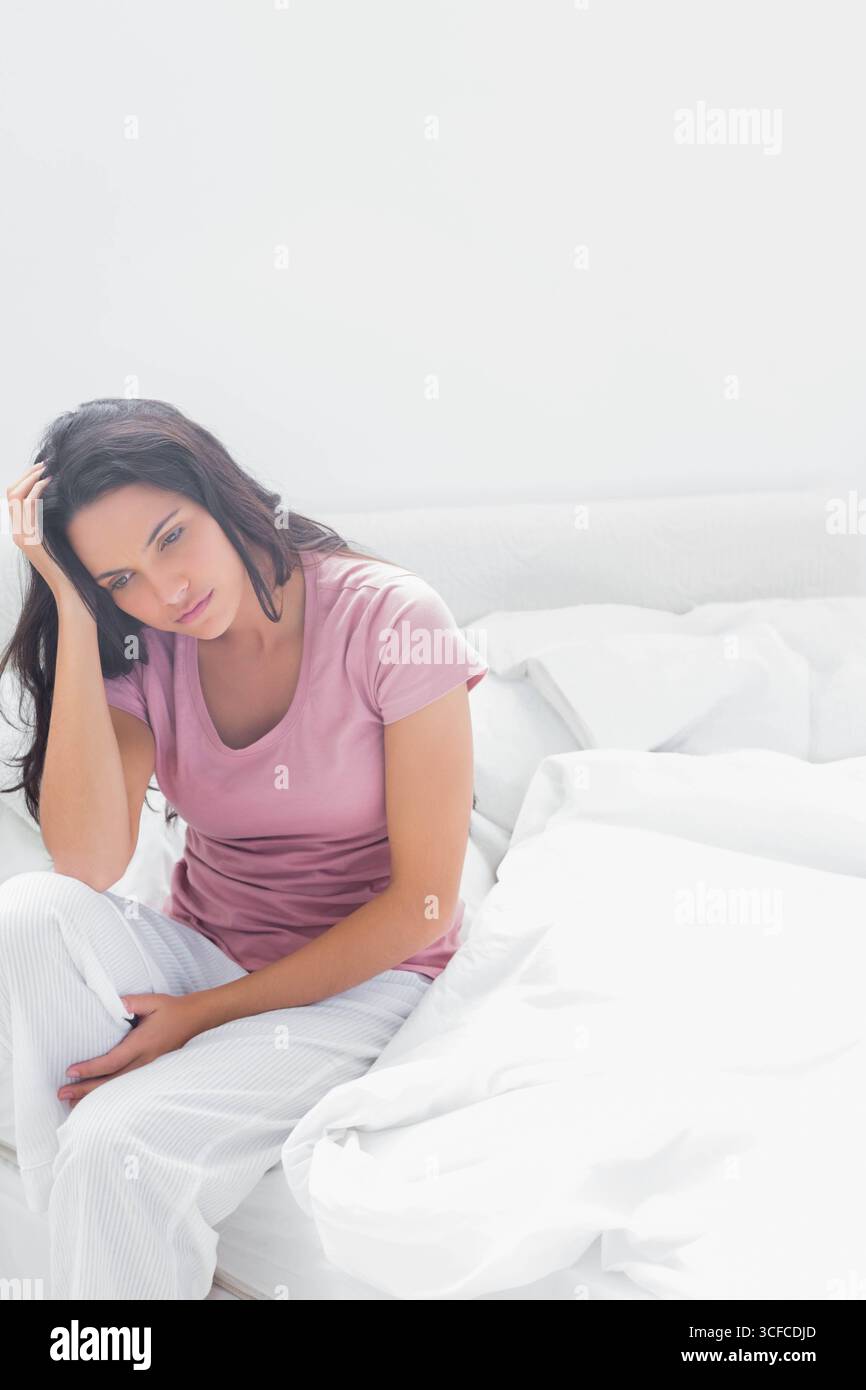 Femme assise sur le lit avec des draps blancs et des oreillers tenant la tête et regardant vers le bas Banque D'Images