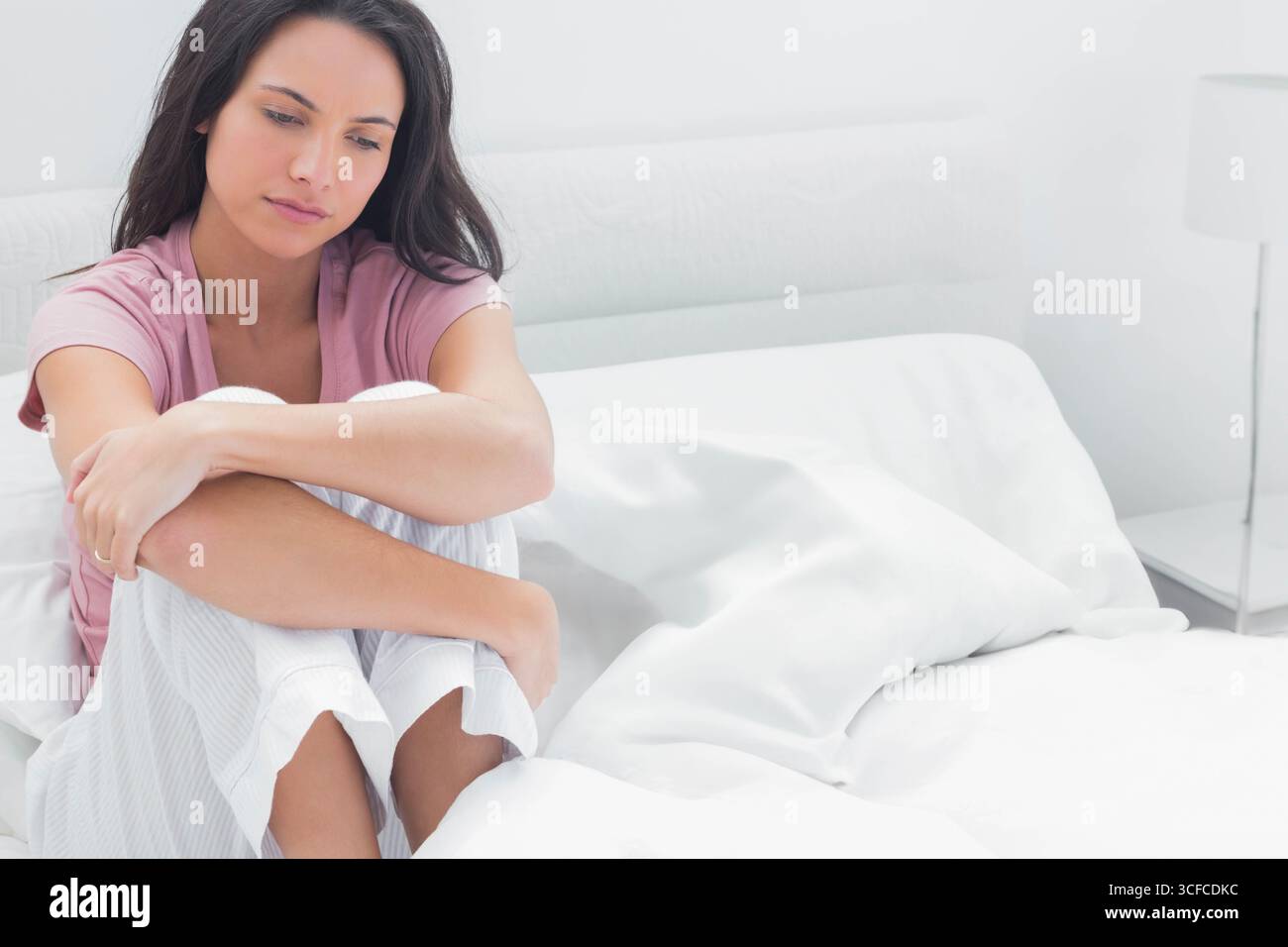 Femme assise en pyjama sur le lit à la maison, regardant vers le bas à la lampe de table de chevet, espace de copie Banque D'Images