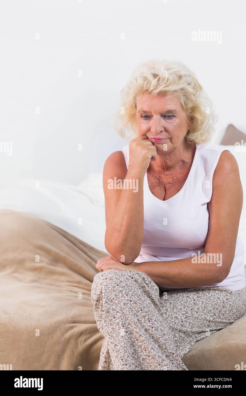 Femme senior assise sur le bord du lit dans une chambre à coucher éclairée regardant la couverture beige, espace copie Banque D'Images