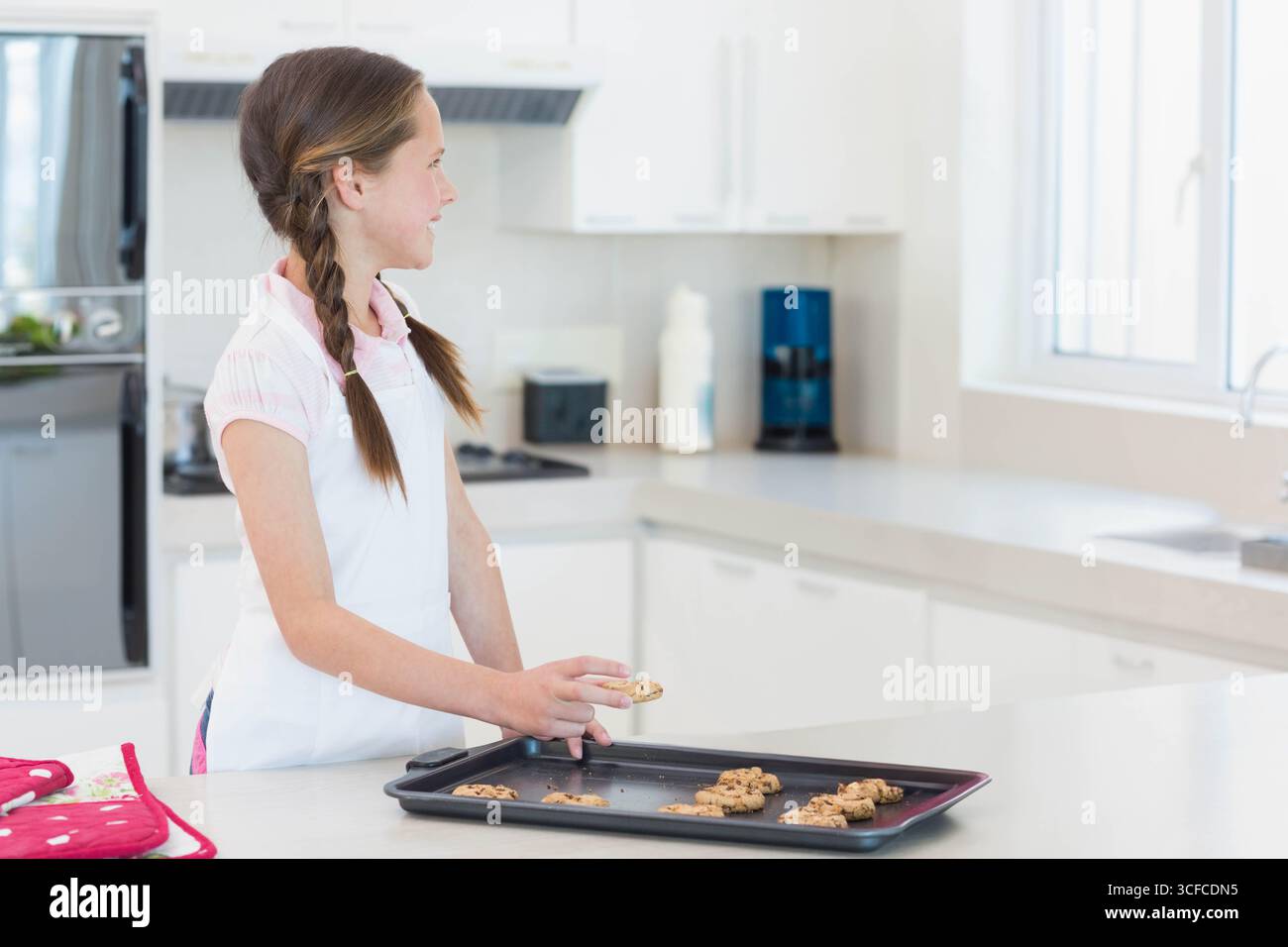 Fille d'âge scolaire dans un tablier arrangeant des biscuits cuits au four sur une feuille de biscuits noire au comptoir de la maison, espace de copie Banque D'Images