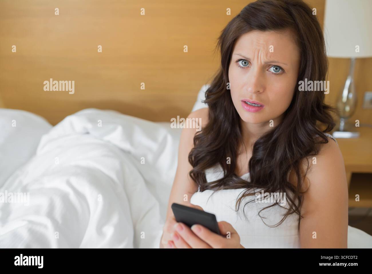 Femme tenant smartphone assis sur le lit avec tête de lit regardant concerné par la lampe sur la table de chevet Banque D'Images