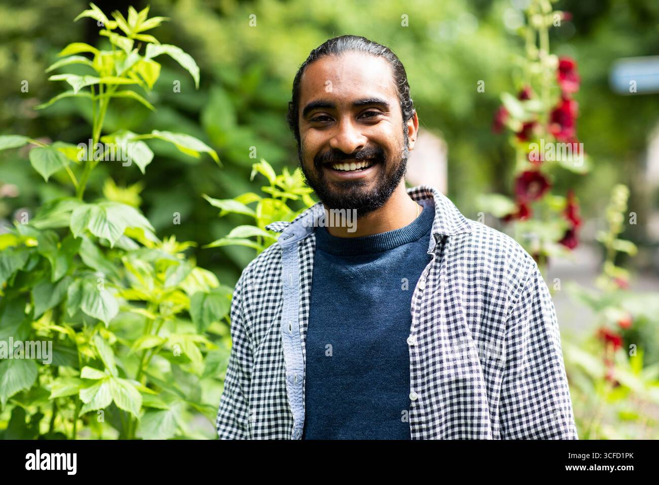 Homme souriant dans un jardin avec des plantes vertes luxuriantes et des fleurs lumineuses en arrière-plan. Allemagne Banque D'Images