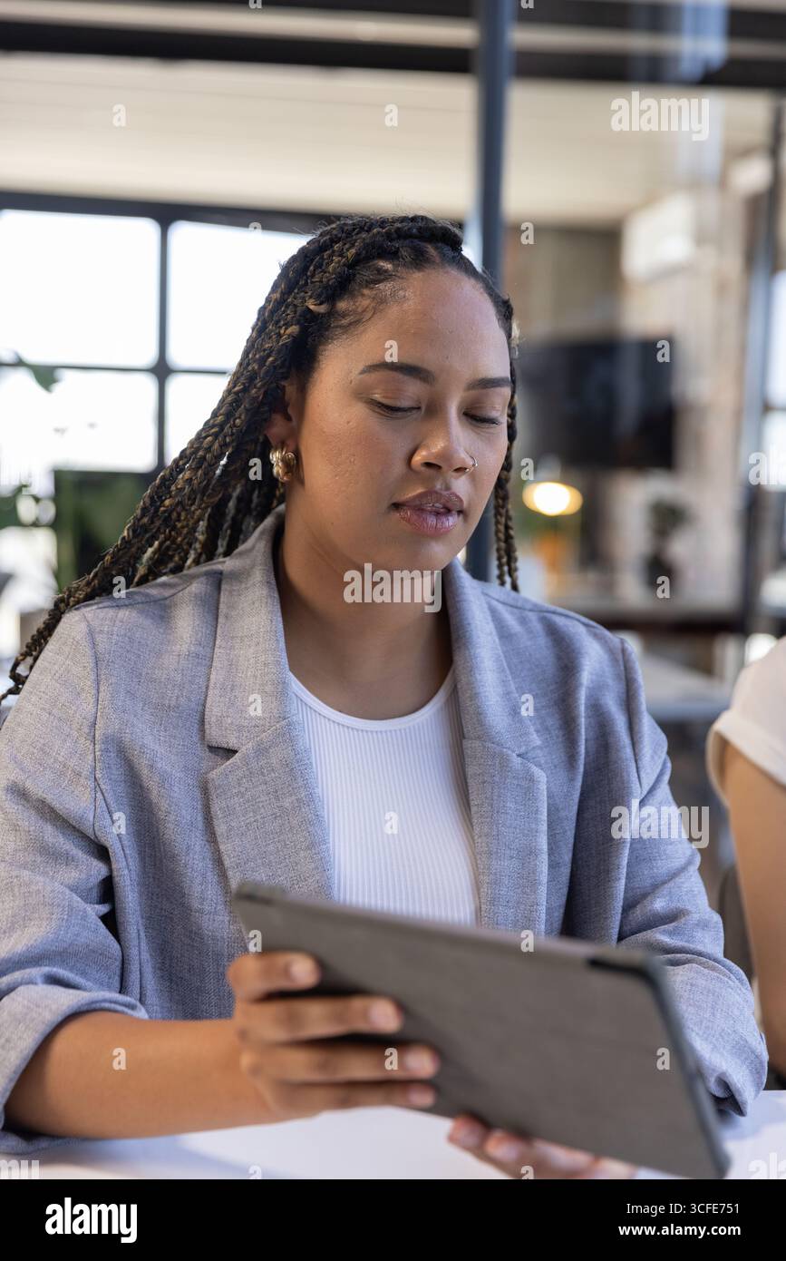 Femme afro-américaine assise au bureau dans le bureau ouvert tenant la tablette tout en regardant les moniteurs Banque D'Images