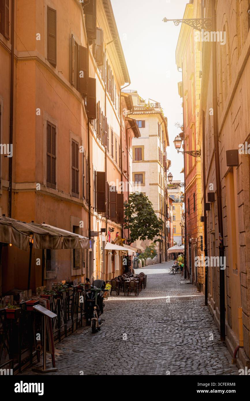Rue pavée étroite à Trastevere, Rome, Italie, avec cafés, tables, lumière chaude du coucher du soleil et bâtiments colorés Banque D'Images