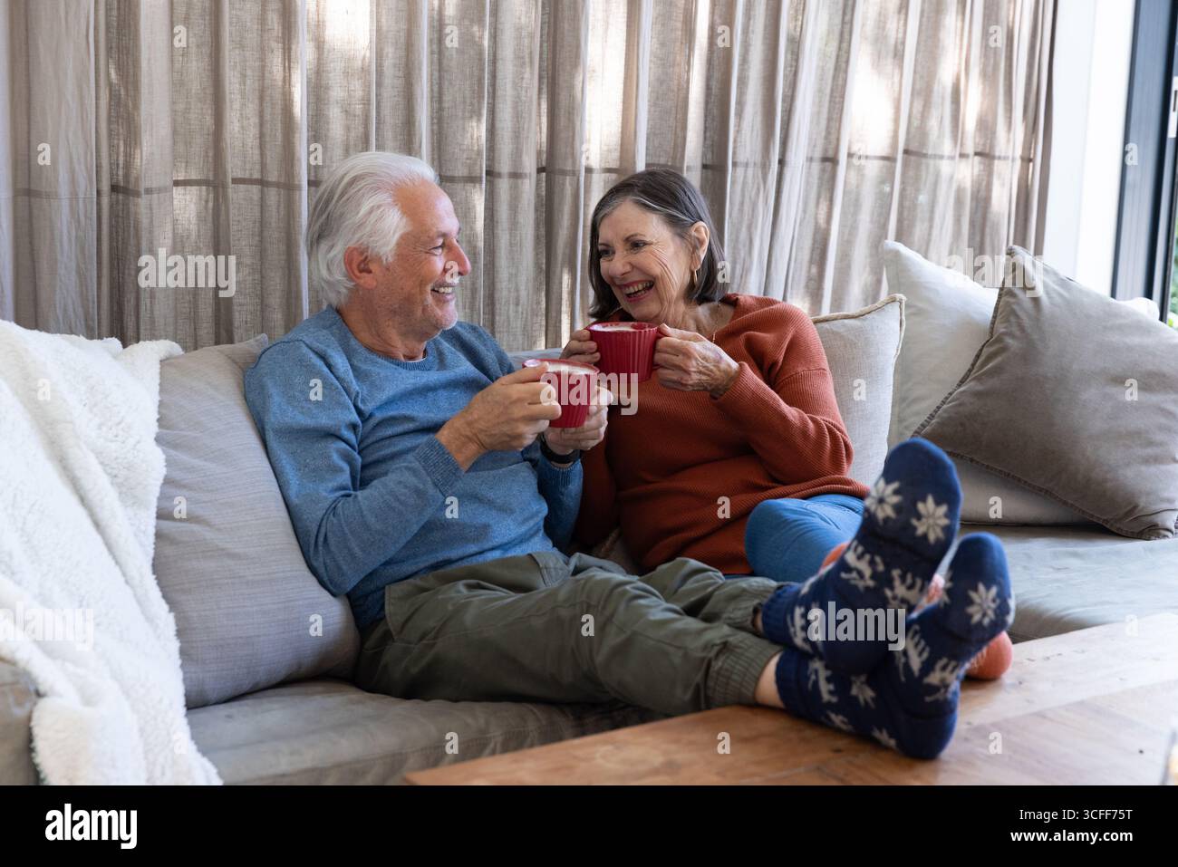Couple senior souriant tout en tenant des tasses rouges sur le canapé de couleur claire dans le salon confortable Banque D'Images