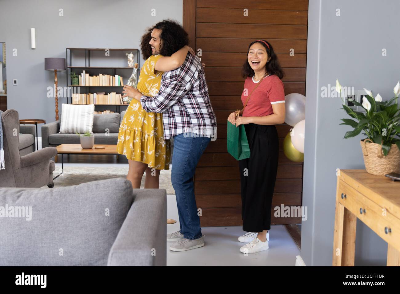 Divers amis embrassant et échangeant sac cadeau vert dans le salon moderne, ballons pastel Banque D'Images