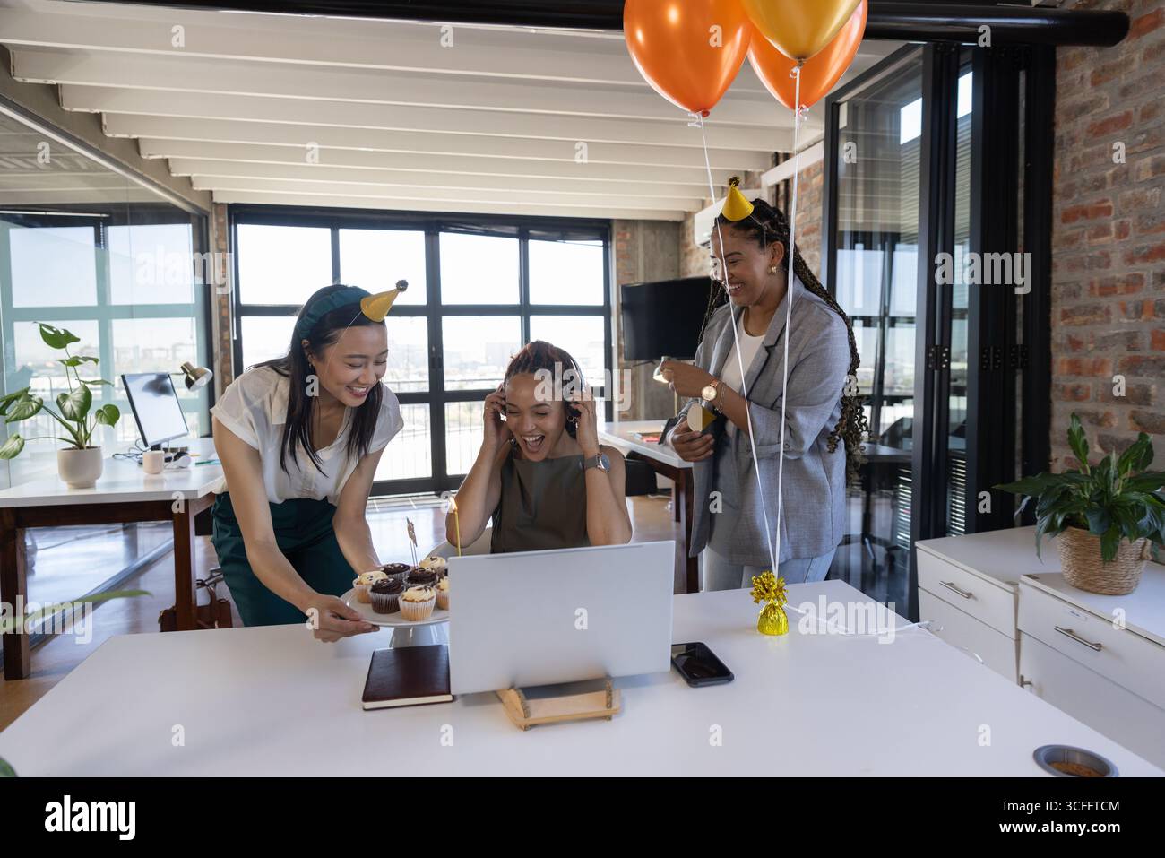 Diverses collègues féminines se réunissant au bureau autour d'un ordinateur portable pour l'anniversaire avec des cupcakes et des ballons Banque D'Images