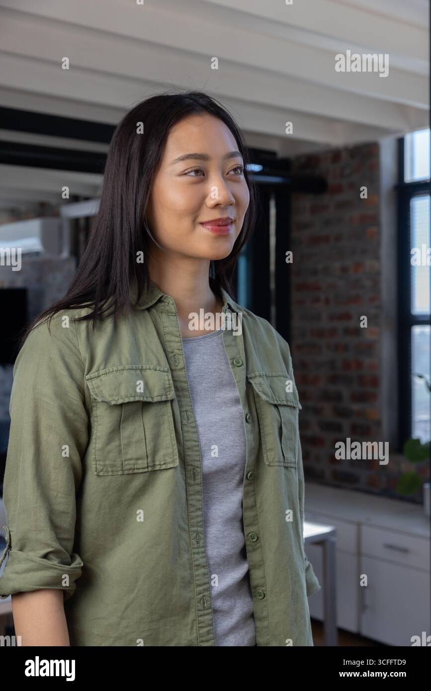 Femme asiatique portant une chemise vert olive debout dans la chambre loft avec moniteur et stores de fenêtre Banque D'Images