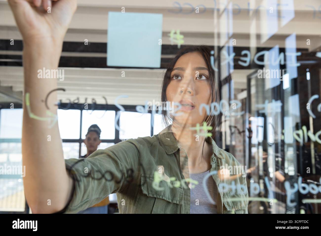 Diverses collègues féminines dessinant des diagrammes sur un tableau blanc en verre avec une note adhésive bleue au bureau Banque D'Images