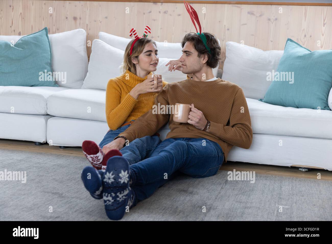 Couple assis à la maison portant des bandeaux en bois et des chaussettes tricotées tenant des tasses en céramique près d'un mur de bois Banque D'Images