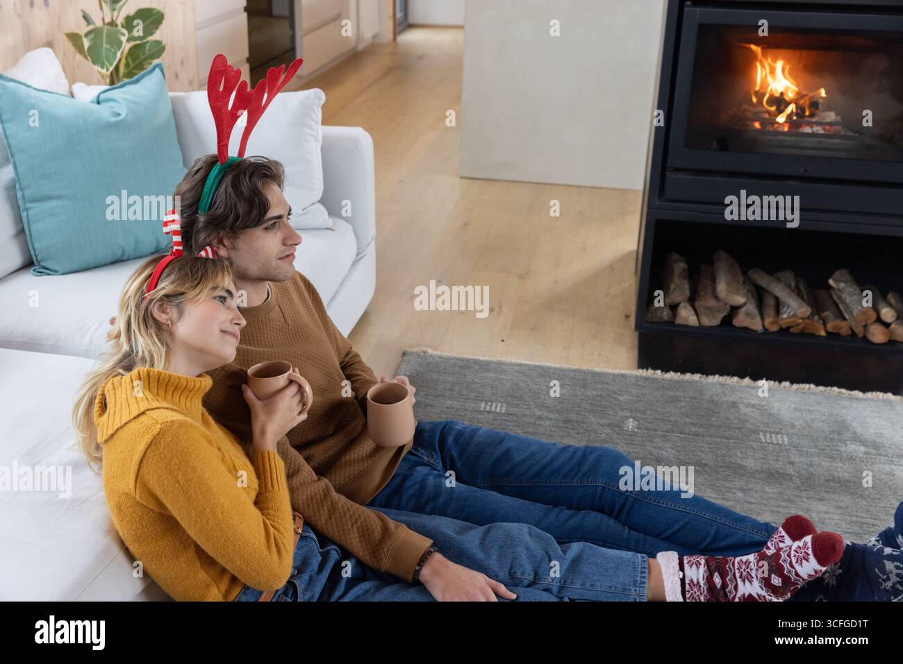 Couple portant des bandeaux en bois rouge appuyé contre le canapé, tenant des tasses à la maison devant la cheminée Banque D'Images