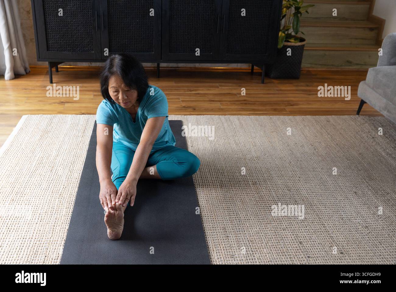 Femme asiatique senior s'étirant en turquoise activewear sur tapis de yoga noir dans le salon, espace de copie Banque D'Images