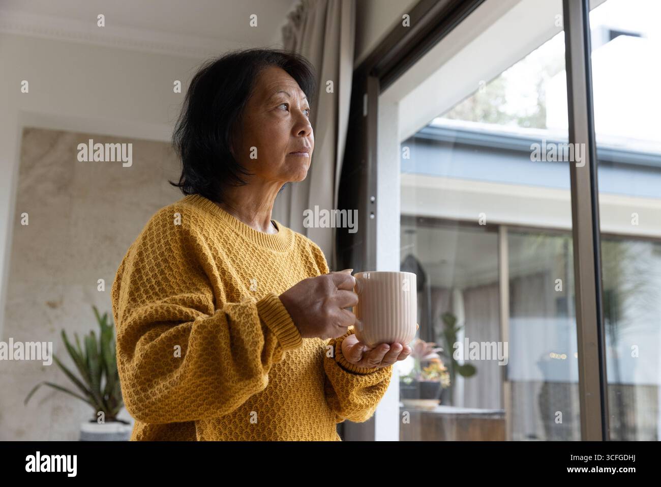 Femme asiatique senior portant un pull moutarde tenant une tasse côtelée regardant à l'extérieur à la porte coulissante en verre Banque D'Images