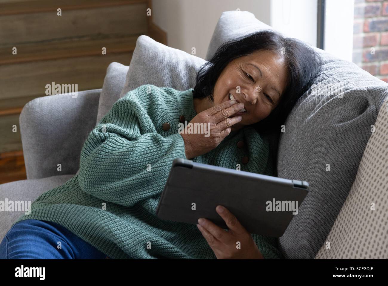 Femme asiatique senior s'inclinant sur le canapé par des escaliers en bois dans le salon à l'aide d'une tablette noire Banque D'Images