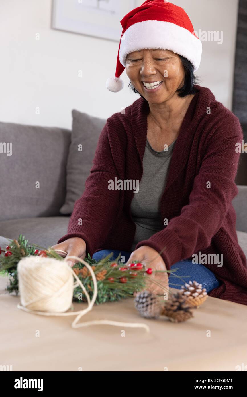 Femme asiatique senior dans le chapeau rouge santa assemblant une couronne avec des pommes de pin et de la ficelle dans le salon Banque D'Images