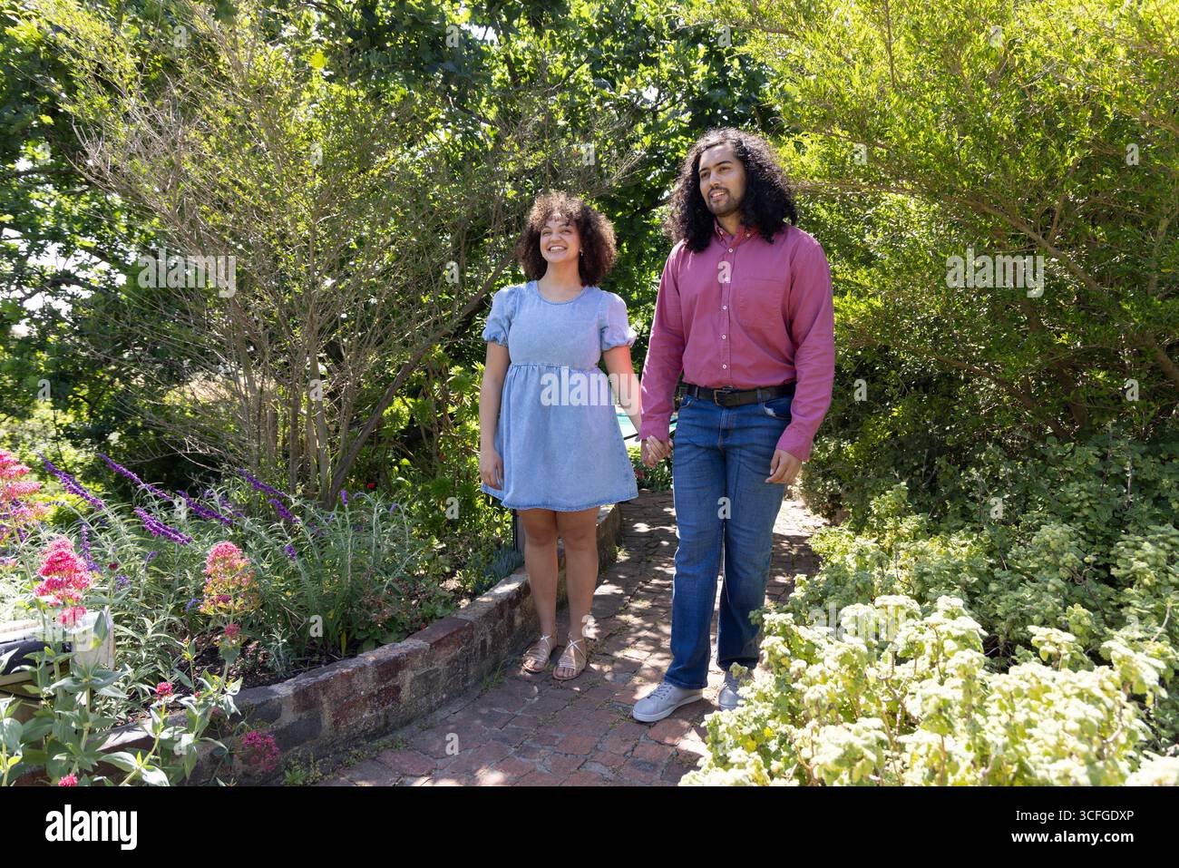 Couple diversifié marchant main dans la main le long du chemin de briques à travers le jardin luxuriant, les plantes à fleurs Banque D'Images