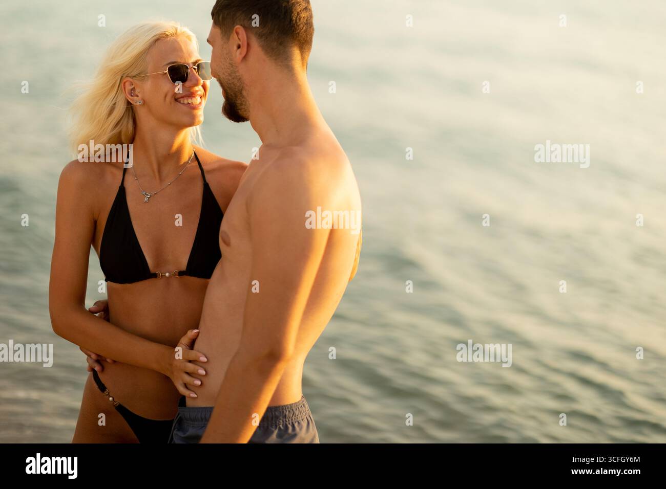 Couple embrassé par le soleil partage des sourires et des moments tendres au bord de l'eau pendant le coucher du soleil, capturant l'essence de la romance estivale et du bonheur. Banque D'Images