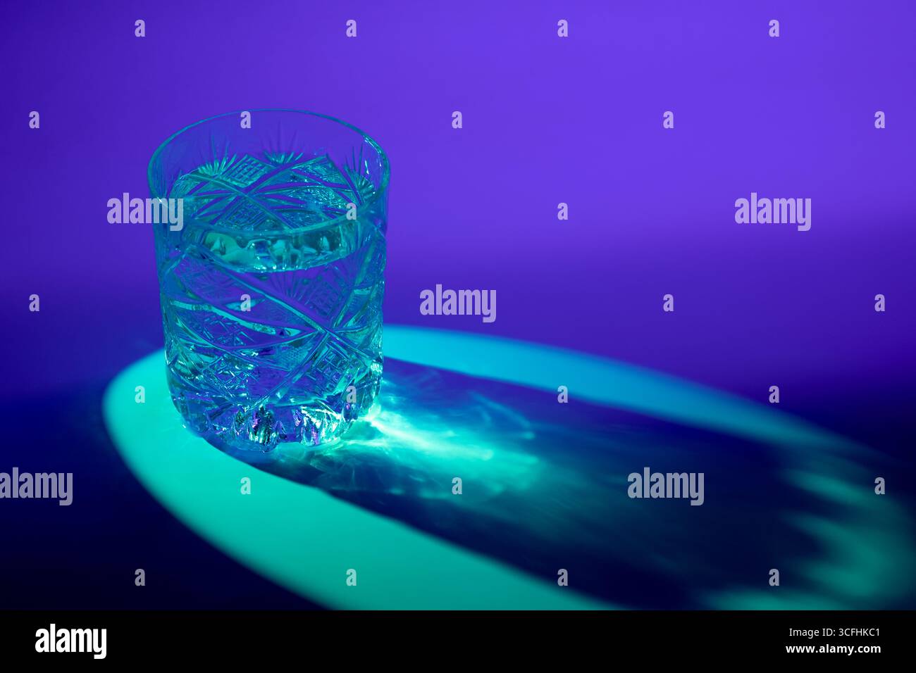 Une photo d'un verre de whisky coupé rempli d'eau, éclairé par un rétro-éclairage bleu, projetant des motifs caustiques sur le sol en raison de sa forme et de sa lumière refr Banque D'Images