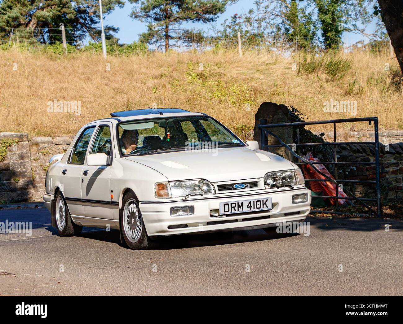 Une voiture Ford Sierra blanche classique conduisant sur une route de campagne ensoleillée avec des arbres et de l'herbe en arrière-plan Banque D'Images