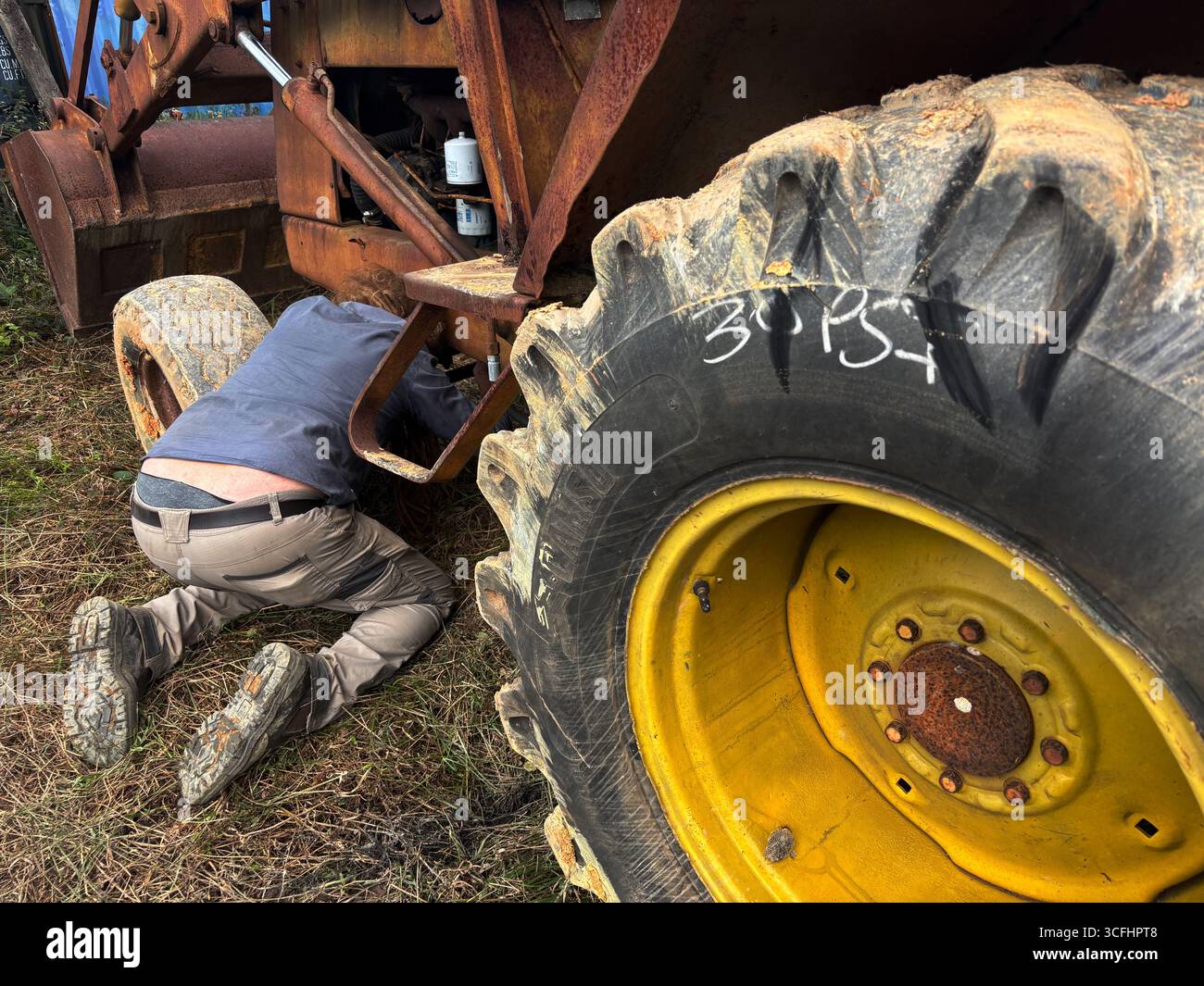 Homme agenouillé sous le châssis du tracteur chargeur dans un environnement agricole boueux. Pas DE MR ou PR Banque D'Images