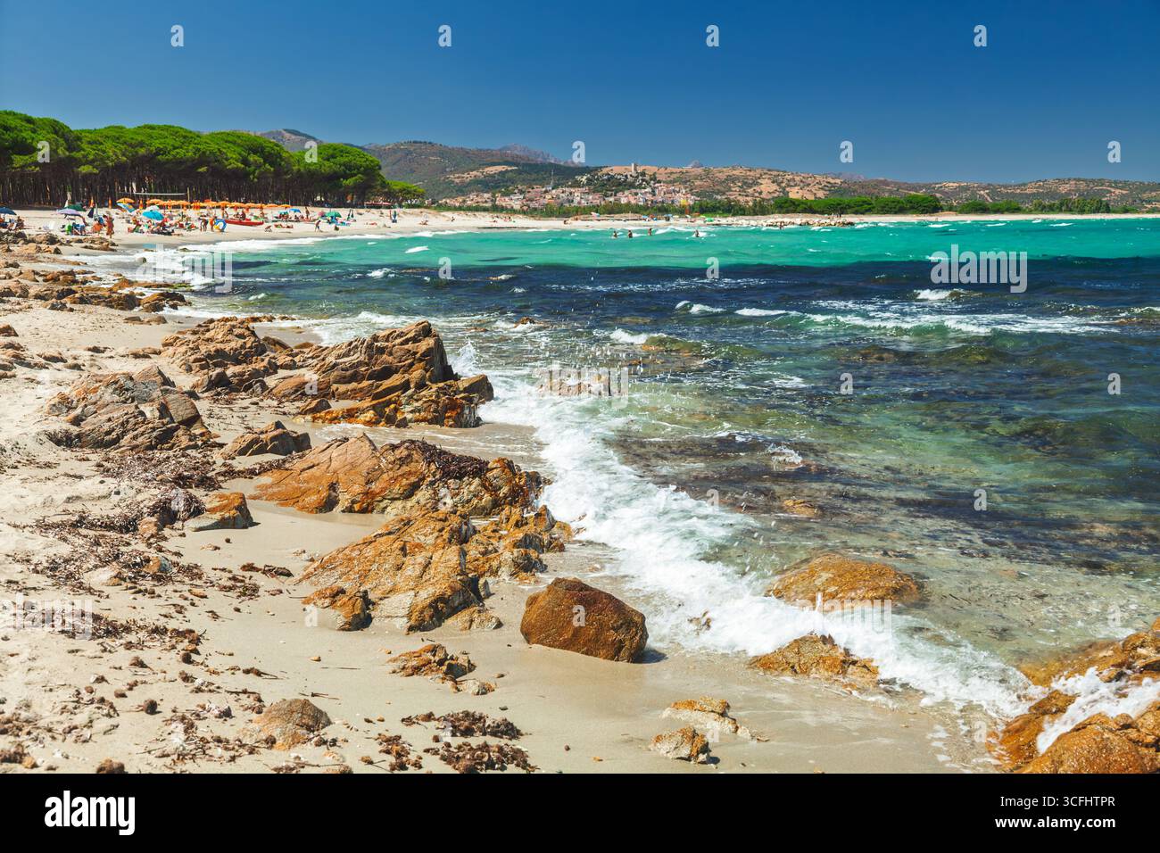 Les touristes profitent d'une journée d'été ensoleillée sur une plage en Sardaigne, Italie, avec des vagues turquoises frappant la rive rocheuse, des pins offrant de l'ombre, et historique Banque D'Images