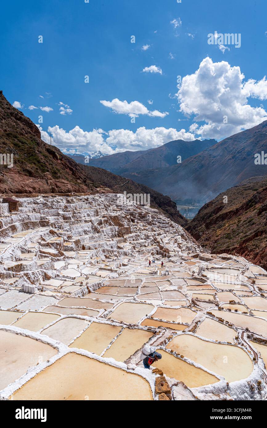 Vue panoramique des mines de sel de Maras à Cusco, Pérou, avec un ouvrier récoltant le sel sous un ciel lumineux. Paysage andin emblématique et patrimoine culturel. Banque D'Images