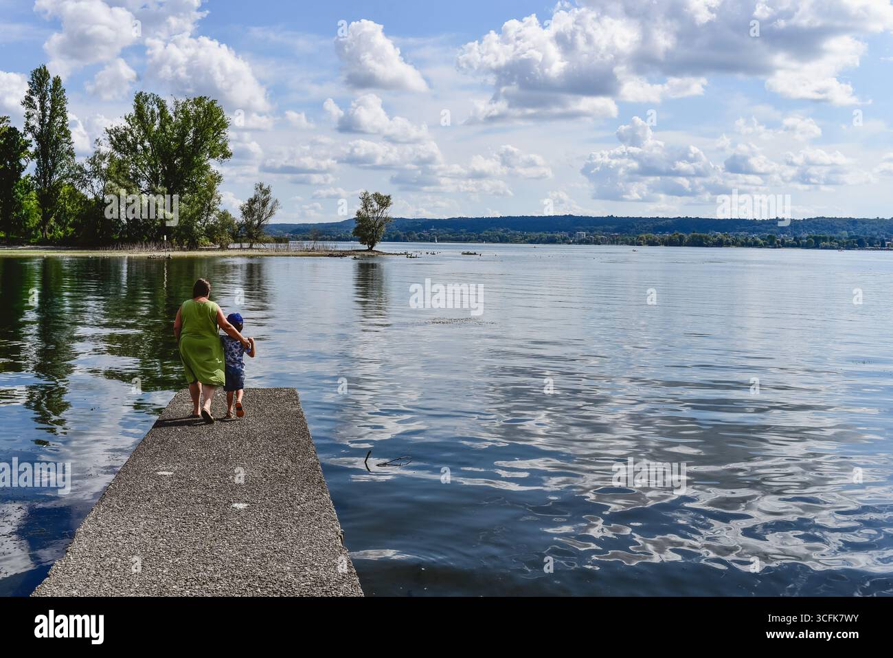 Une mère et son fils se promènent main dans la main le long d'une jetée paisible du lac sous un ciel dégagé avec des nuages moelleux. Banque D'Images