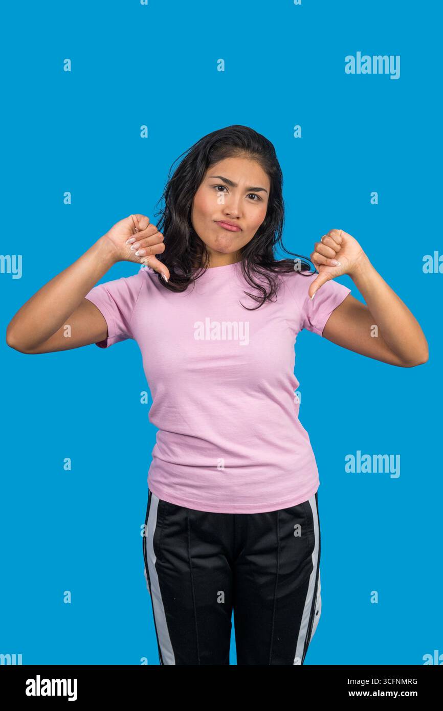 Jeune femme montrant la désapprobation avec le pouce vers le bas geste, exprimant l'aversion et la rétroaction négative sur fond bleu Banque D'Images