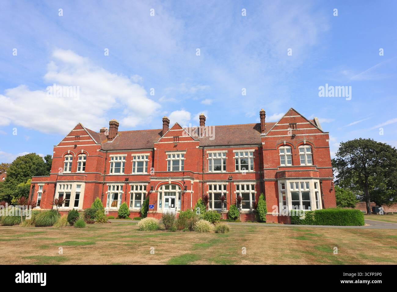 Ce bâtiment en briques rouges bien préservé de l'ancien site de l'hôpital Haslar présente des fenêtres à garniture blanche, un toit à pignons et un design symétrique. Probablement un ancien dortoir ou bloc administratif, il se trouve au milieu d'une pelouse avec des zones sèches et des arbustes dispersés. La scène mélange l'architecture institutionnelle classique avec des signes de changement saisonnier sous un ciel lumineux de septembre. Illustration, image éditoriale, visite de l'ancien site de l'hôpital Haslar à Gosport, Hampshire, Angleterre. 22 août 2025. Banque D'Images