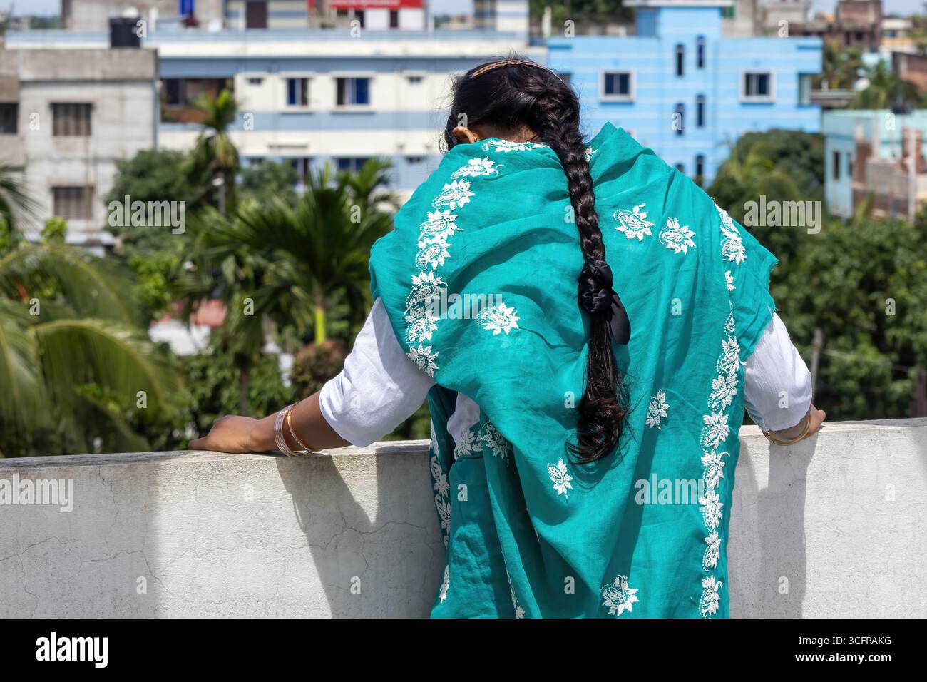 Femme bengali dans un saree turquoise, ses longs cheveux tressés visibles de l'arrière, se tient sur un toit regardant vers le bas. Banque D'Images