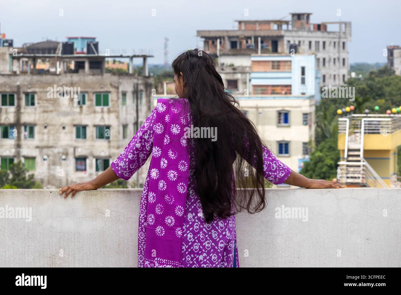Vue arrière d'une belle femme bangladaise sur un toit avec de longs cheveux noirs, portant un kameez salwar en coton batik violet, admirant la vue sur la ville. Banque D'Images