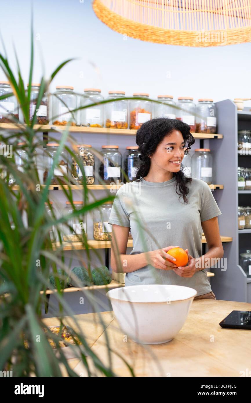 Jeune vendeuse tenant une orange dans un magasin zéro déchet avec des produits alimentaires en vrac Banque D'Images