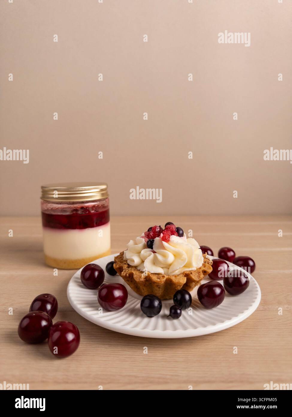 Une petite tarte avec de la crème fouettée et des baies sur une assiette blanche, entourée de cerises. En arrière-plan, un pot de dessert en couches avec des fruits, de la crème, un Banque D'Images