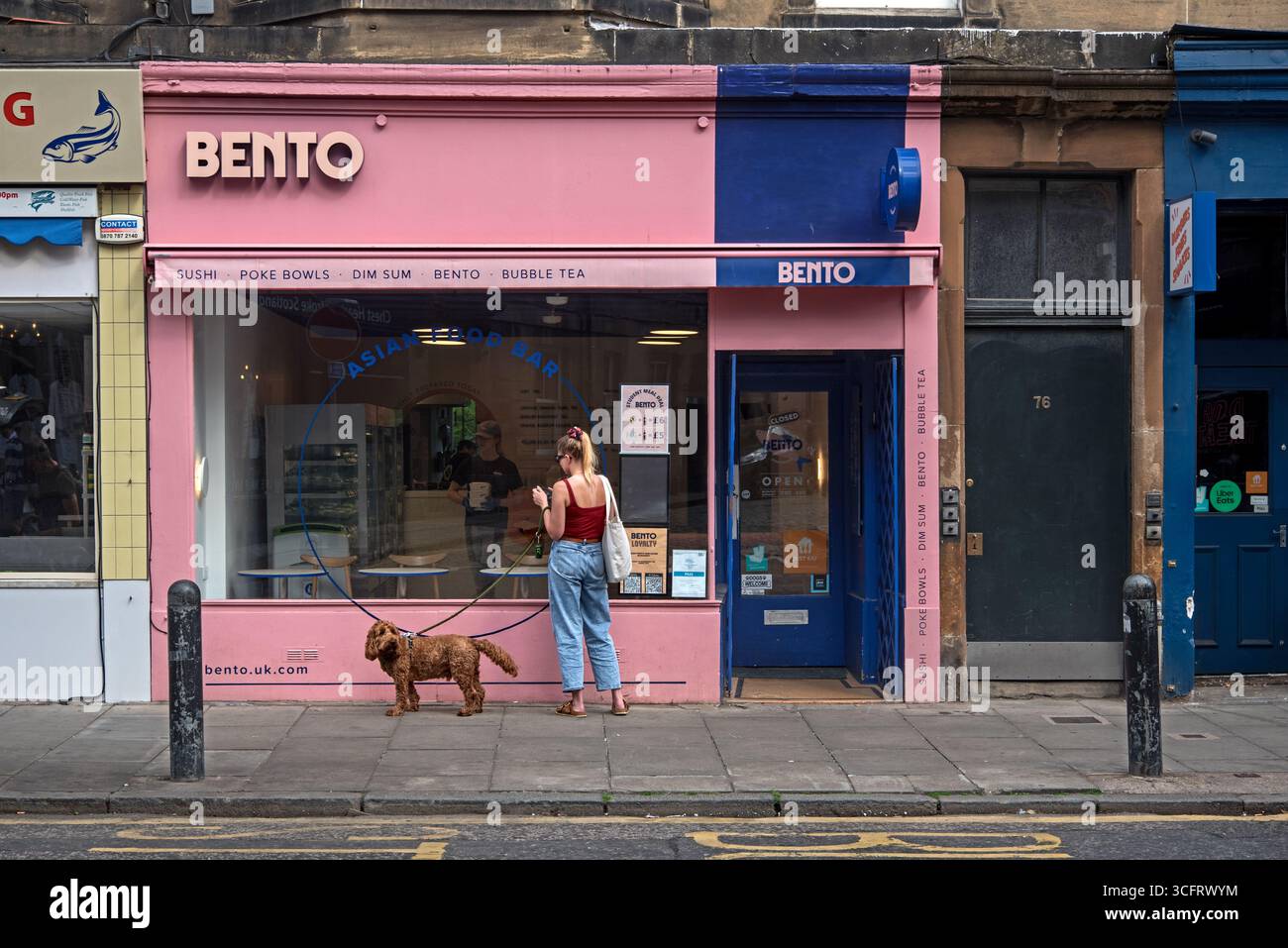 Jeune femme et chien debout devant le bar alimentaire asiatique Bento à Raeburn place, Stockbridge, Édimbourg, Écosse, Royaume-Uni. Banque D'Images