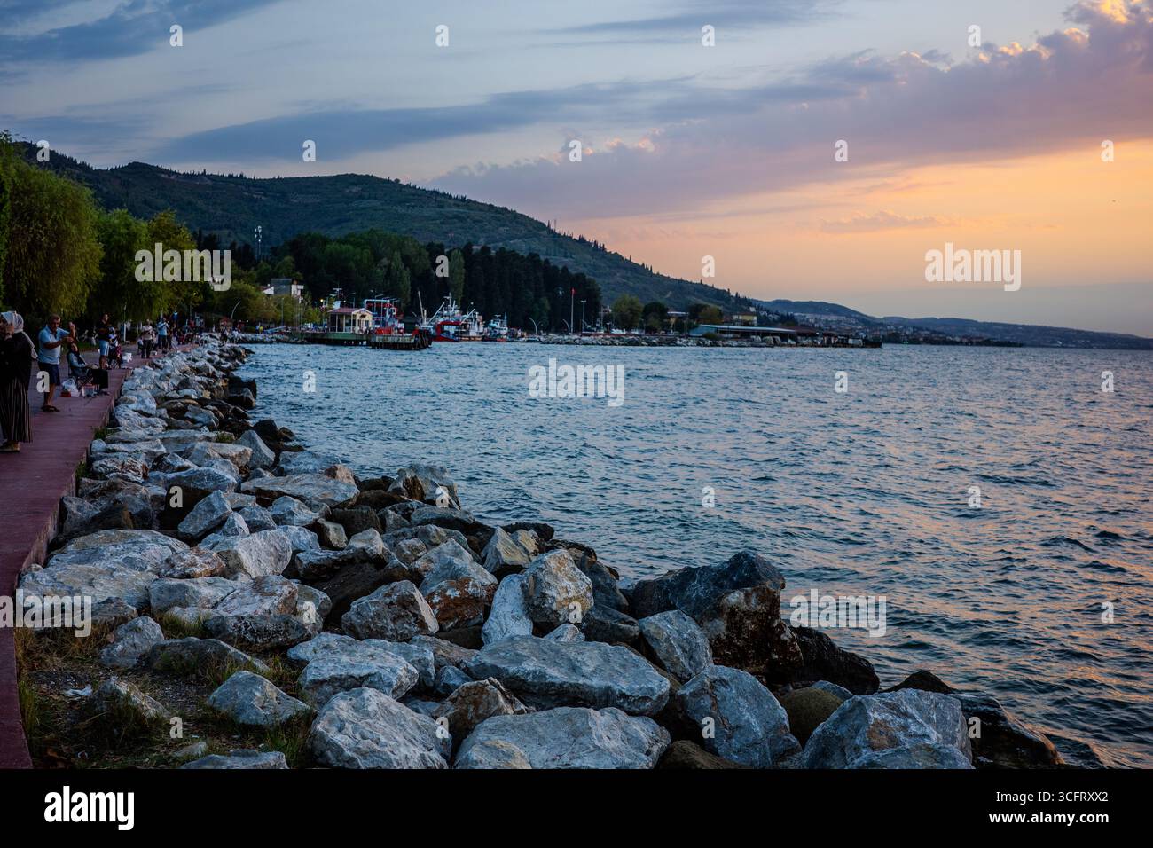 Les gens marchent le long de la promenade à côté de la rive rocheuse de la mer de Marmara pendant le coucher du soleil à Karamürsel, Kocaeli, Türkiye, avec des bateaux et des collines vis Banque D'Images