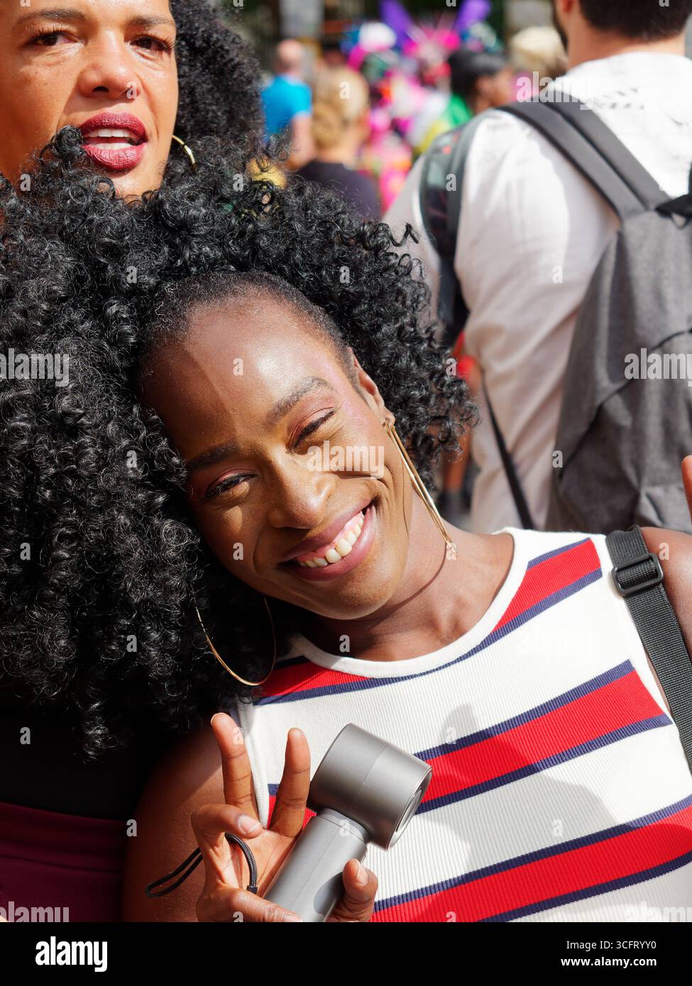 Jolie femme noire avec haut rayé sourit largement tout en donnant le signe de la paix pendant le Carnaval de Notting Hill, Londres. Angleterre 25 août 2025 Banque D'Images