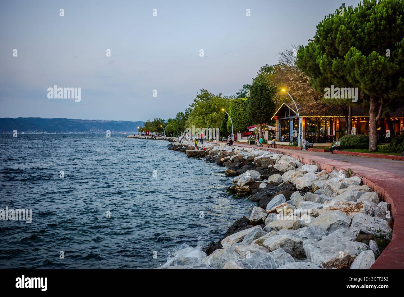 Une promenade au bord de l'eau avec des cafés et des arbres borde le rivage rocheux de la mer de Marmara à Karamürsel, Kocaeli, Türkiye, pendant la soirée. Karamürs Banque D'Images