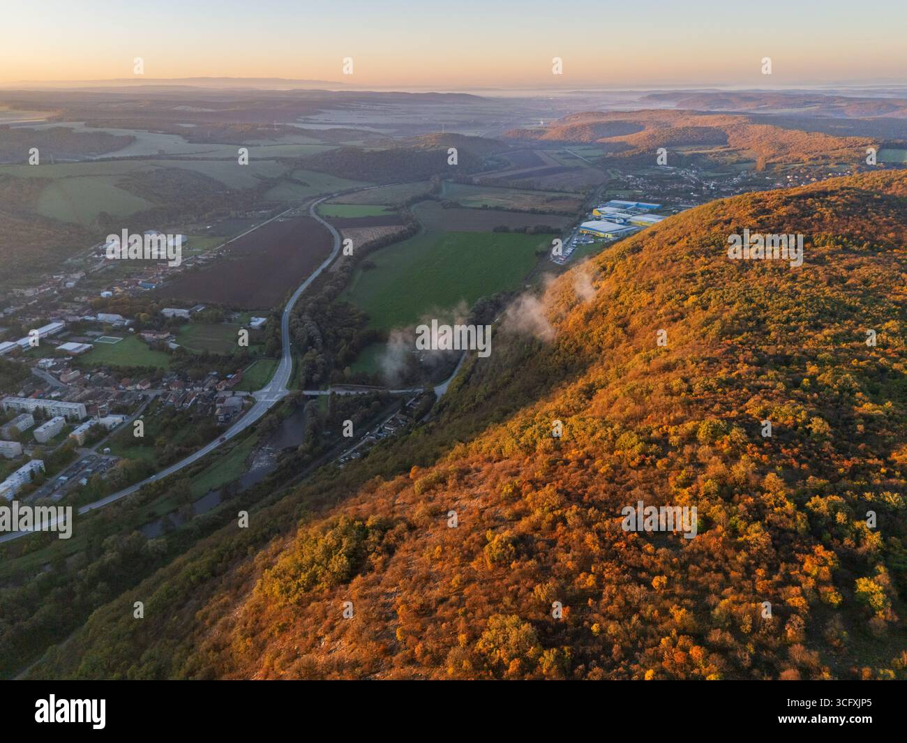 Vue aérienne de la lumière dorée du soleil embrassant les collines escarpées, une vallée sereine qui se déroule en contrebas, et la ville nichée paisiblement, Plešivec, région de Košice, Slovaquie. Banque D'Images