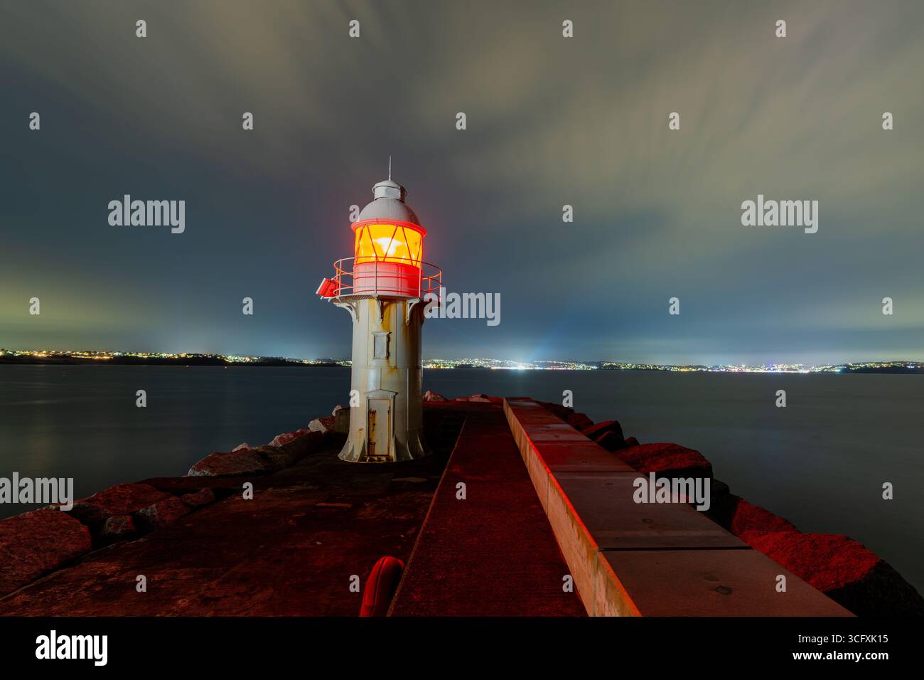 Lumière rouge d'un phare guidant les navires la nuit, situé sur une jetée rocheuse, avec des lumières de la ville visibles au loin Banque D'Images