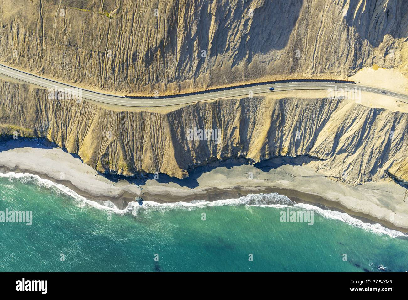 Vue aérienne d'une route sinueuse serpente le long de la côte accidentée, où les falaises embrassées par le soleil rencontrent la mer turquoise, créant un contraste étonnant de c Banque D'Images