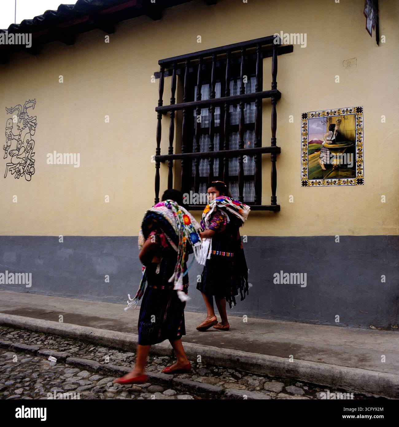 Zwei Frauen mit bunten Textilien gehen auf einem Gehweg vor einem Gebäude mit Wandbild und vergittertem Fenster vorbei. Guatemala, um 1980. Banque D'Images