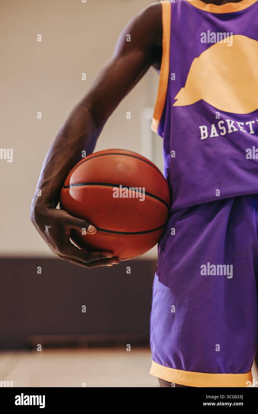 Gros plan d'un athlète portant un uniforme violet et or tenant un basket-ball en milieu de match, montrant détermination et concentration. Set de terrain de sport intérieur Banque D'Images