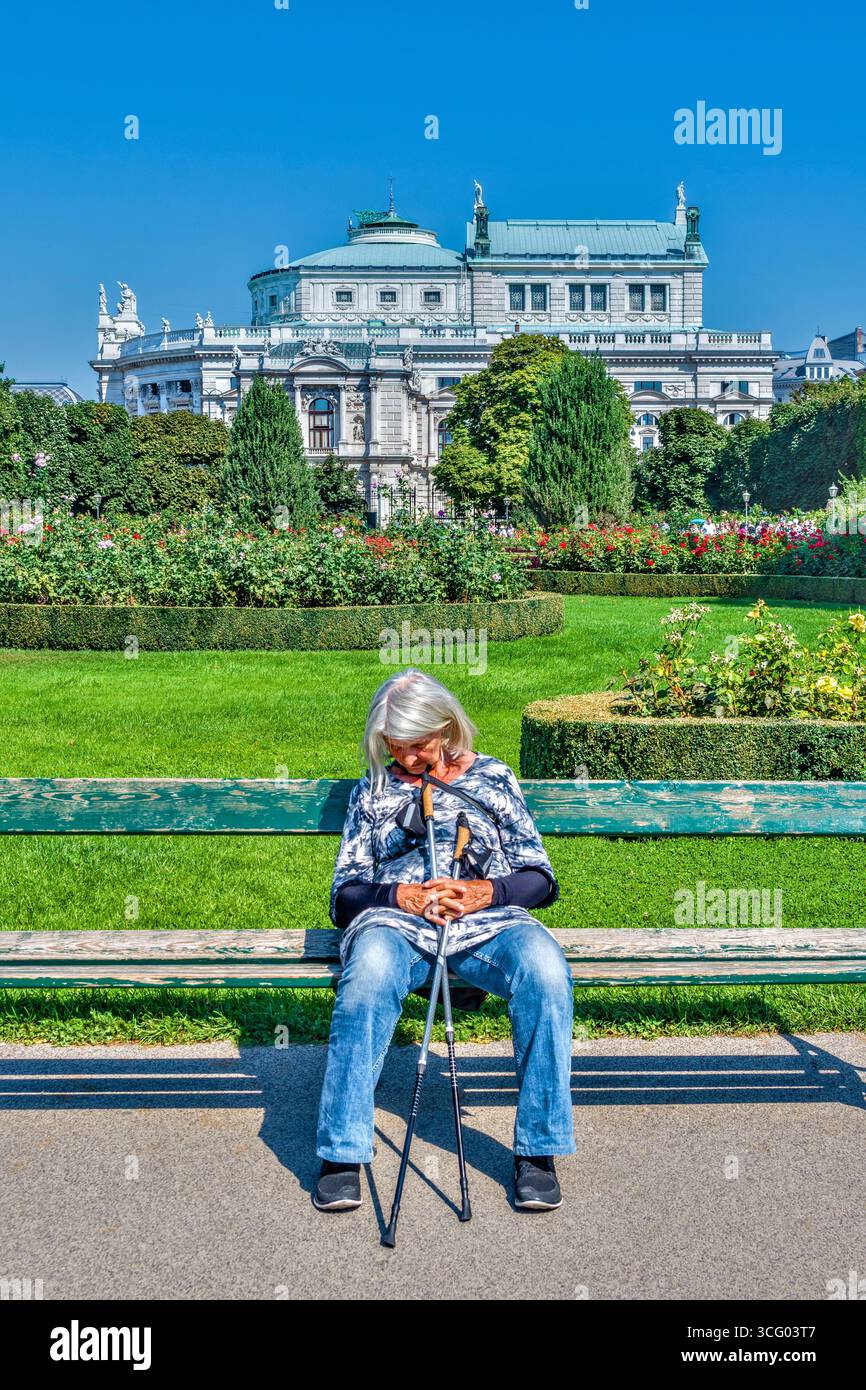 Femme âgée avec des cannes de marche reposant sur le banc du parc dans le Volksgarten (jardin du peuple) avec le Burgtheater en arrière-plan - Vienne, Autriche. Banque D'Images