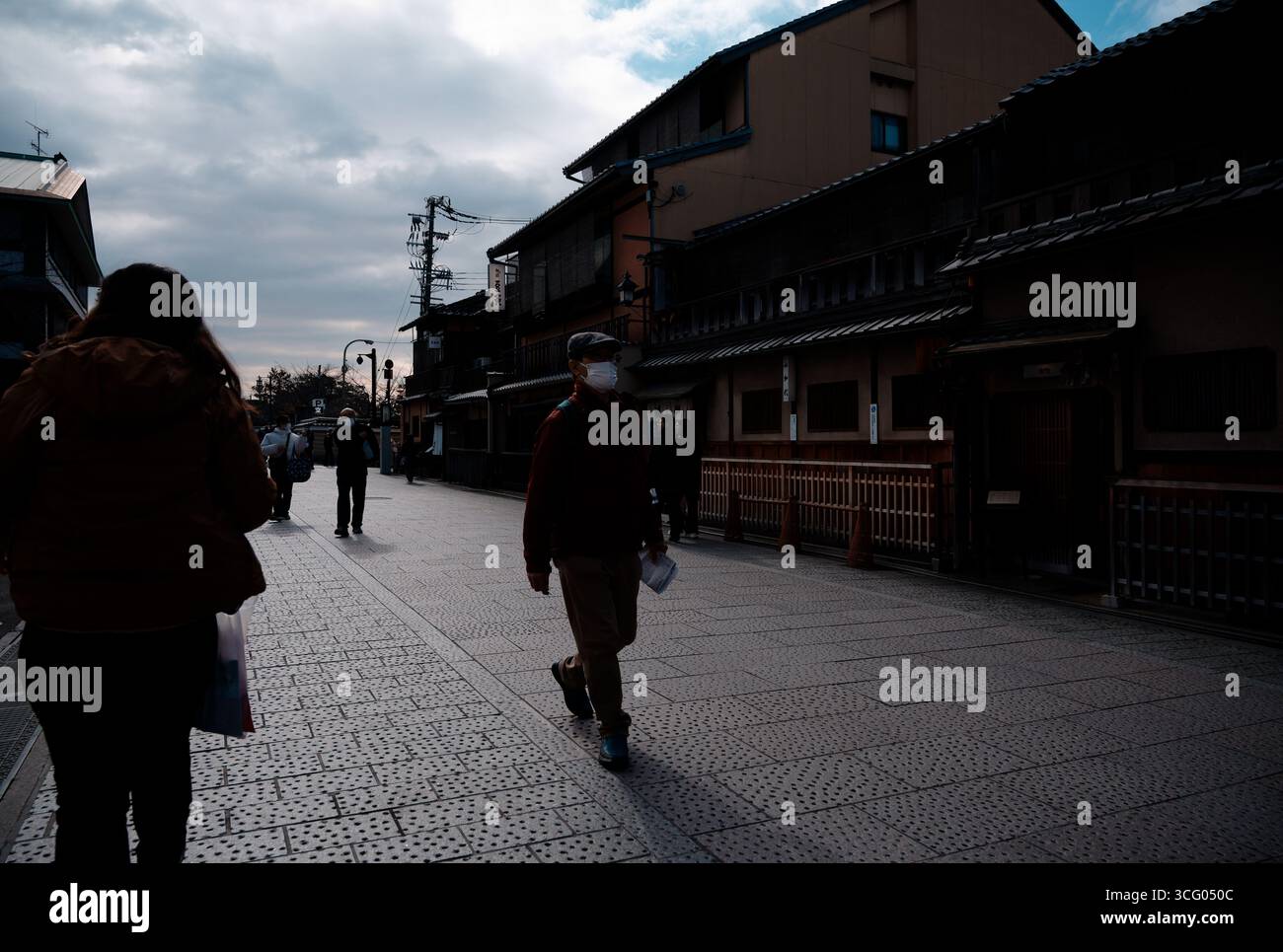 Figure masquée marchant le long d'une rue traditionnelle de Kyoto au crépuscule, projetant de longues ombres sous un ciel d'hiver Banque D'Images
