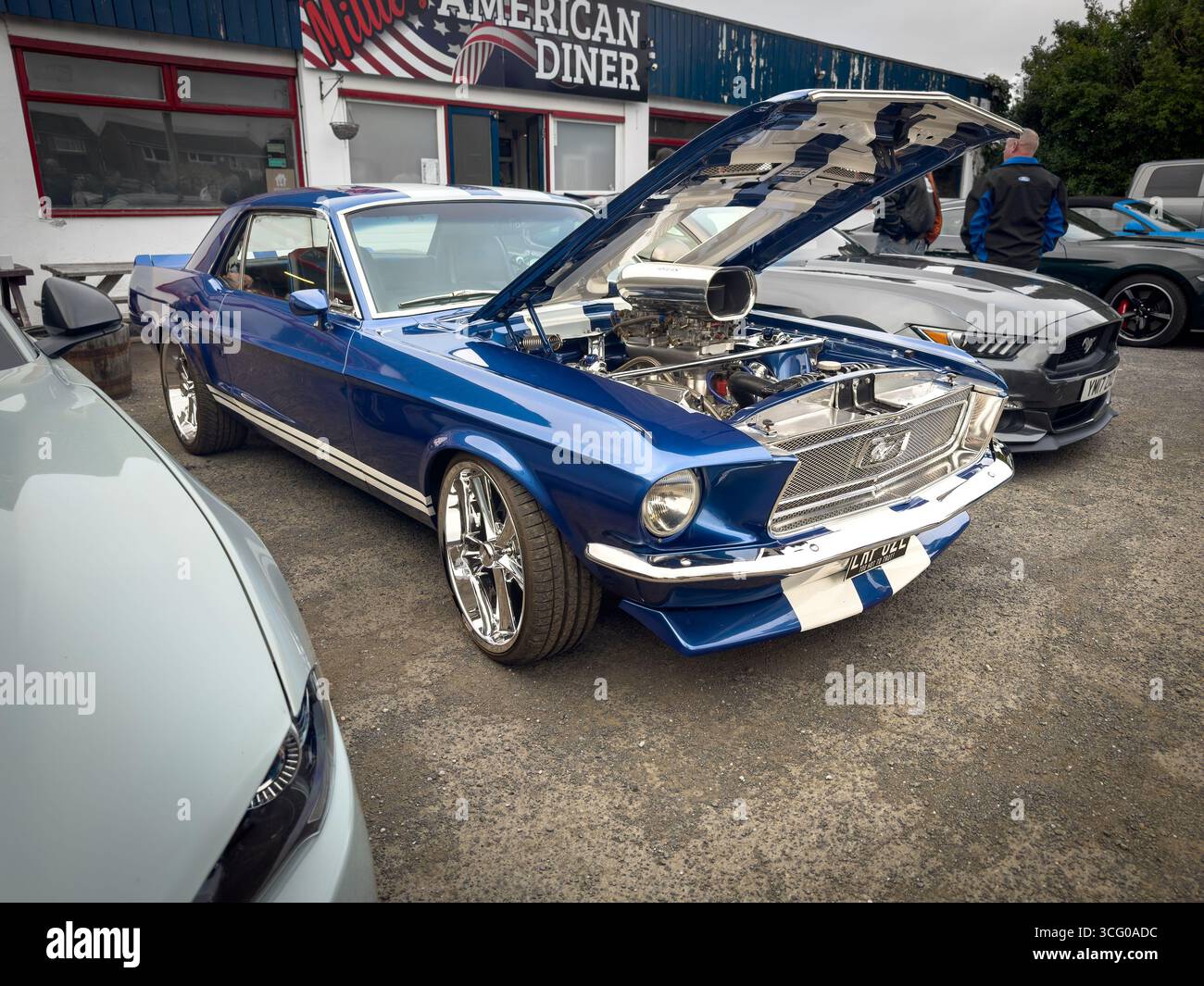 Une Ford Mustang bleue classique avec son capot ouvert montrant son moteur garé devant un restaurant américain parmi les voitures modernes - Image de stock capturée avec un smartphone