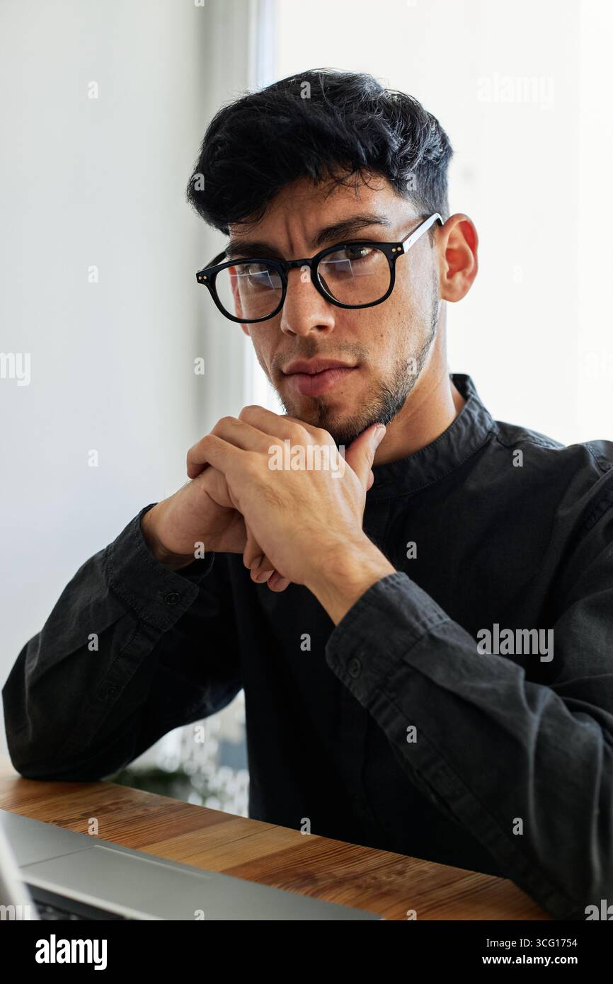 Portrait vertical d'un jeune homme hispanique pensif assis au bureau dans le bureau reposant son menton sur les mains regardant la caméra Banque D'Images