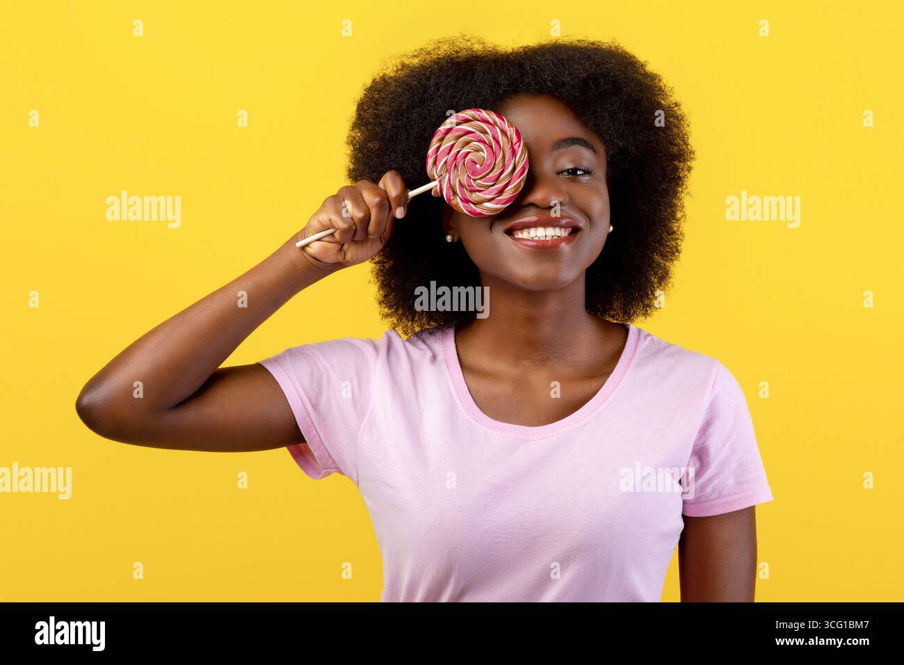 Dent douce. Jeune femme afro-américaine positive couvrant un œil avec un lollipop sur fond jaune vif de studio Banque D'Images